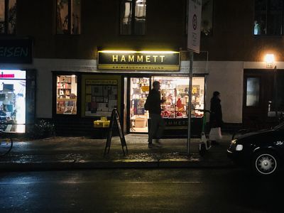 Nachtaufnahme der Hammett-Buchhandlung von Außen. Logo in Gelb und Schwarz, zwei erleuchtete Fensterscheiben. Dahinter sind Bücher zu erkennen.