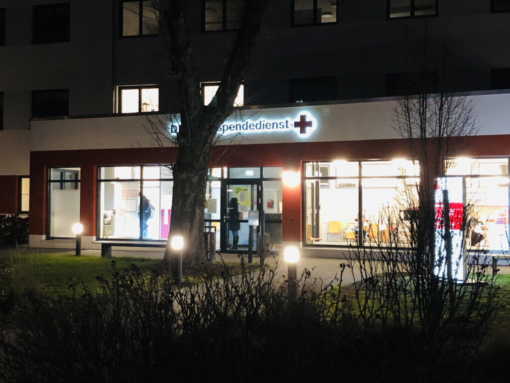 Foto von außen auf den beleuchtetenden Blutspendedienst. Im Inneren sind Stühle und wartende Menschen erkennbar.