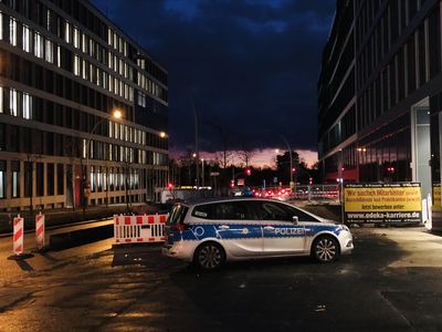 Polizeiwagen steht zwischen zwei Bürohäusern. Spätabends, es hat geregnet, künstlicher Lichter spiegeln sich, im Hintergrund ein Sonnenuntergang.
