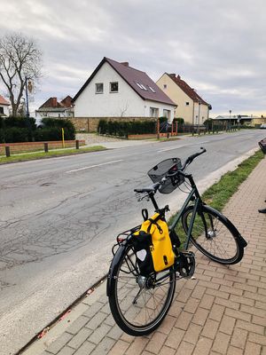 Fahrrad mit Fahrradtasche und Lenkerkorb steht auf einem Fußweg. Daneben eine Dorfstraße in schlechtem Zustand, im Hintergrund zwei Einfamilienhäuser.