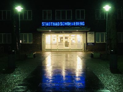 Blick auf den Eingang des Stadtbad Schöneberg. Es ist nacht, hat geregnet, auf dem Nassen Boden spiegeln sich blaue und grüne LIchter.
