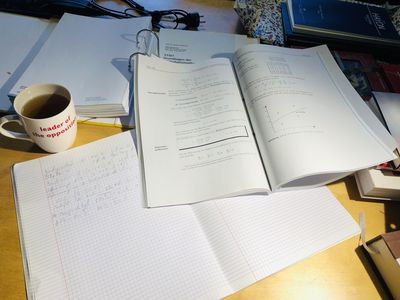 Schreibtischaufsicht mit aufgeschlagenem Heft (Einträge mit Bleistift) und Mathe-Kurs-Unterlagen. Danaben eine Tasse Tee.
