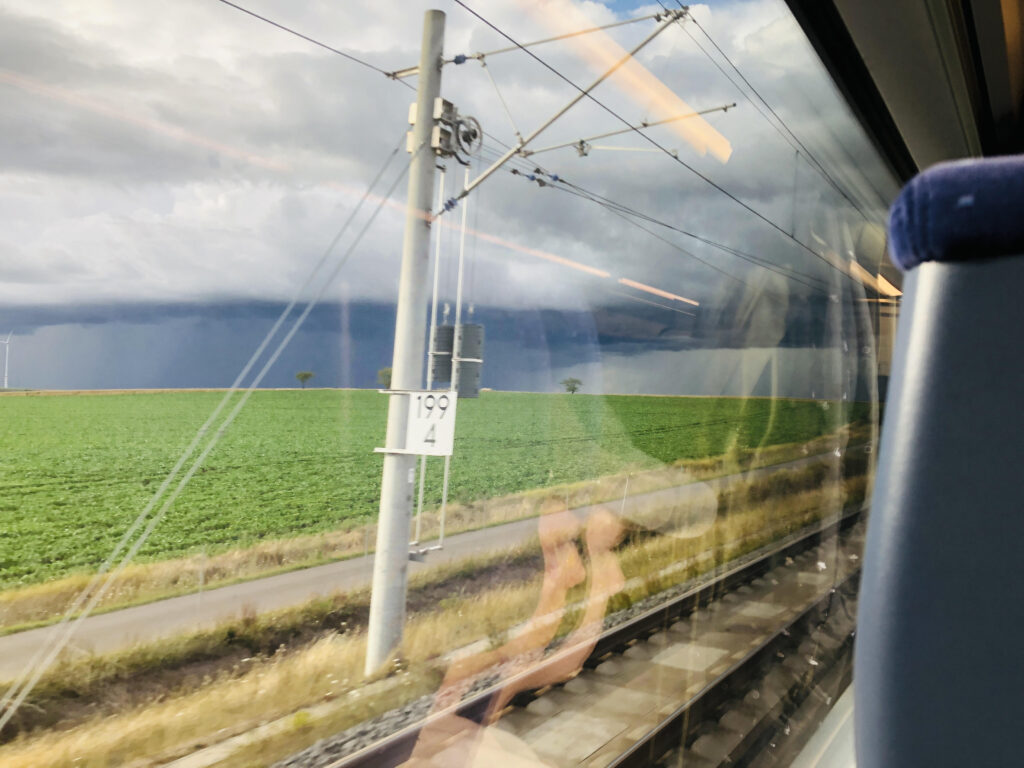 Blick aus dem Zugfenster. Strommast, grüner Acker im Hintergrund Unwetterwolken.