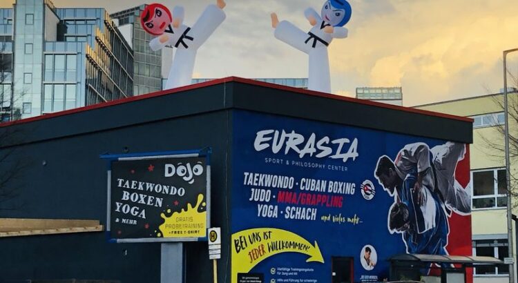 Werkstattgebäude, in dem nun ein Dojo Werbung für Taekwondo, MMA, Yoga und Schach macht. Dramatischer Sonnenuntergangshimmel, auf dem Dojo zwei Aufblasfiguren, die Karateka darstellen sollen.