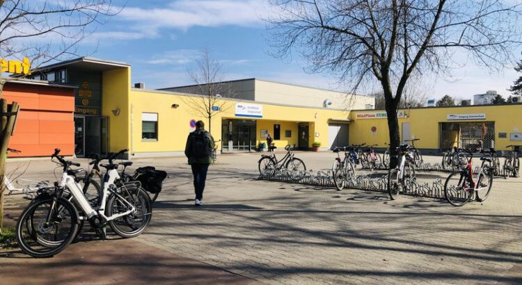 Blick auf den Vorplatz Kombibad Gropiusstadt vor strahlend blauem Frühlingshimel. Ein noch kahler Baum, angeschlossene Fahrräder, ein Mann der auf das Bad zuläuft.