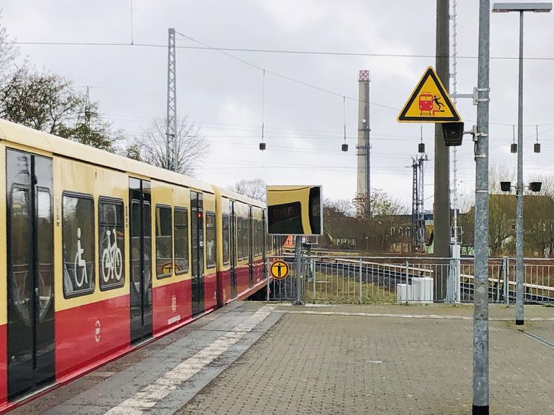 Blick vom Bahnhof Hennigsdorf auf den Rest des Bahnsteigs, eine einfahrende S-Bahn, einen Industrieturm und mehrere Signal- und Leitungsmasten.