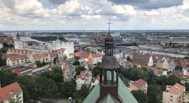 Blick vom Turm der Jakobikirche. Im Vordergrund Kirchendach mit Laterne, im Hintergrund Hafen und Altstadt. Wilde Wolken am Himmel.
