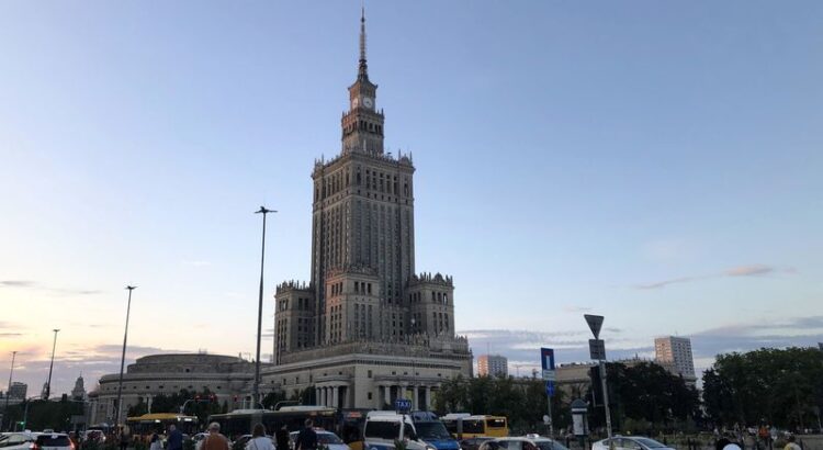 Warschauer Kulturpalast in Abendstimmung. Im Vordergrund Verkehr und laufende Menschen.