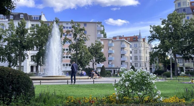 Blick auf den Viktoria-Luise-Platz Berlin: Grünfläche, Fontäne, im Vordergrund ein Blumenbeet, im Hintergrund eine Mischung aus Altbau und Nachkriegsneubau. Blauer Himmel mit einzelnen Wolken.