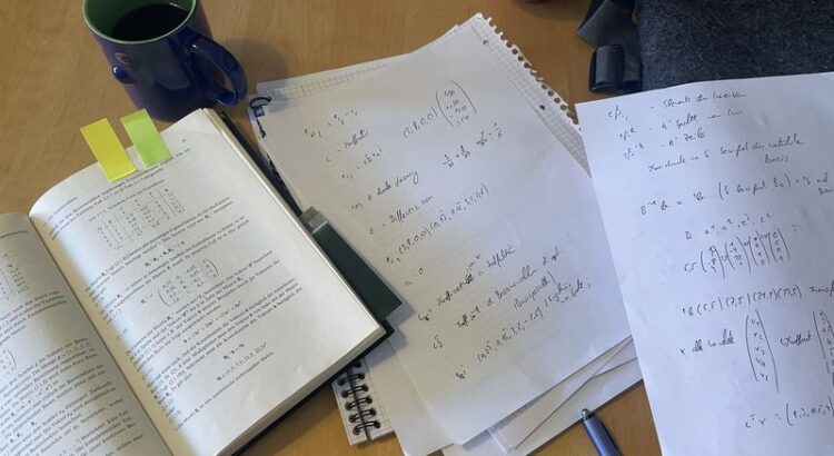 Blick auf den Schreibtisch. Ein aufgeschlagenes Buch Wirtschaftsmathematoik von 1973, zwei Zetteln mit Notizen dazu und eine Kaffeetasse und ein Stift.