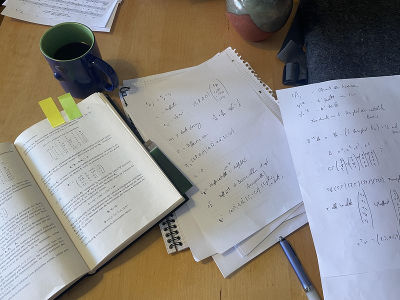 Blick auf den Schreibtisch. Ein aufgeschlagenes Buch Wirtschaftsmathematoik von 1973, zwei Zetteln mit Notizen dazu und eine Kaffeetasse und ein Stift.
