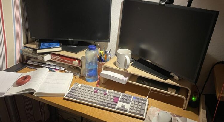 Bild vom Schreibtisch mit zwei ausgeschalteten Monitoren, Tastatur, Maus, Mousepad, zwei Schreibtischaufsätzen - drumherum diverse Bibliotheksbücher zu mathematischer Programmierung, Stifte und ein Taschenrechner.