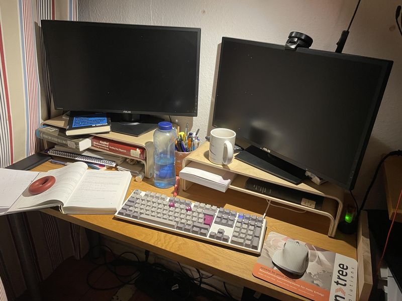 Bild vom Schreibtisch mit zwei ausgeschalteten Monitoren, Tastatur, Maus, Mousepad, zwei Schreibtischaufsätzen - drumherum diverse Bibliotheksbücher zu mathematischer Programmierung, Stifte und ein Taschenrechner.
