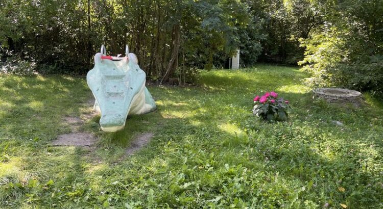 Ein Stück grüner Wiese vor Büschen. Darauf links eine ehemalige Miniaturrutsche in Form eines Krokodils, erkennbar in die Jahre gekommen und mit Flatterband zum Rutschen gesperrt, rechts ein Tuff rot blühender Blumen.