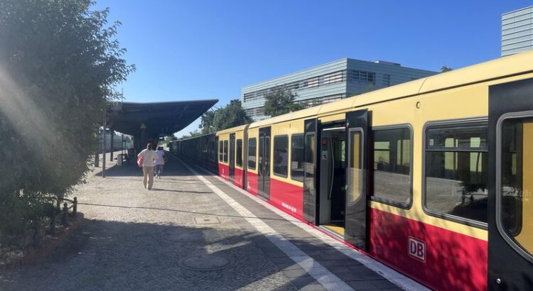 Unüberdachter S-Bahn-Bahnsteig, mit S-Bahn auf der rechten Seite. Strahlend blauer Himmel, im Hintergrund zu erahnen ein grünes modernes Bürogebäude.