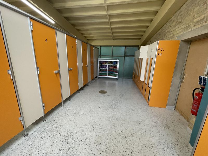 Blick in einen Schwimmbad-Kabinentrakt mit orangen Türen der Umkleidekabinen (links) und orange-beigen Spinden (rechts)- Am Ende des Gangs: Ein Käfig mit Schwimmnudeln etc.