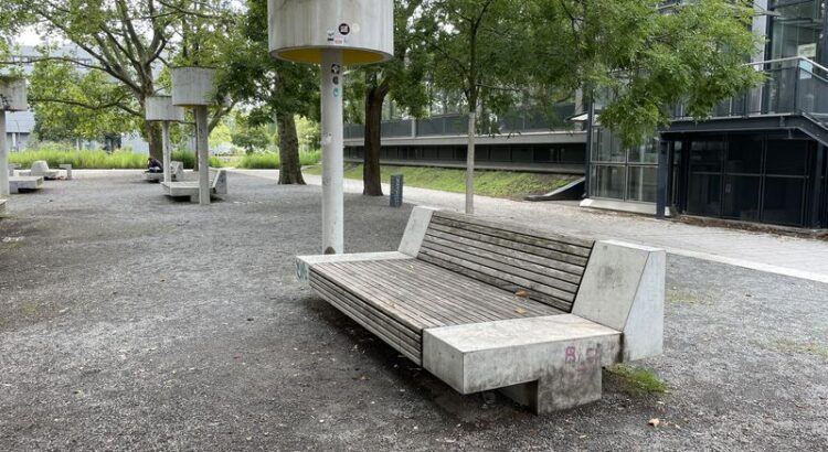 Kiesplatz mit mehreren leeren Sitzgelegenheiten, die eine Mischung aus Bank und Liegestühl sind)., Darüber Bäume, und eine Art Säulen. Alles zusammen eher brutalisch in der Anmutung,