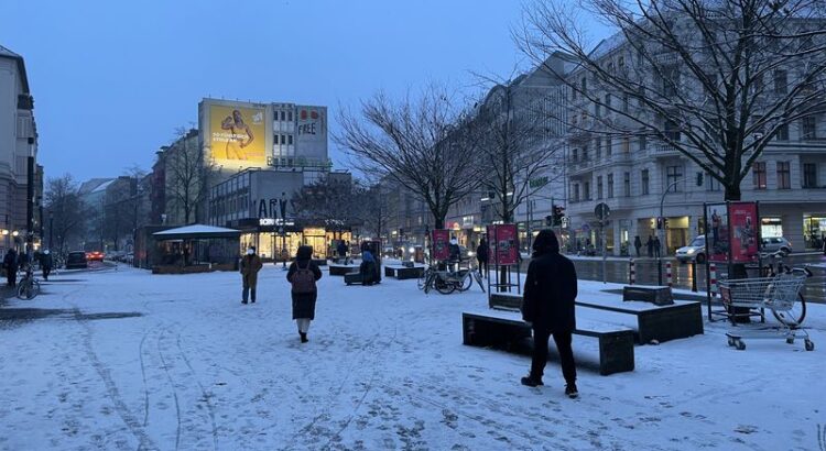 Alfred-Scholz-Platz Neukölln. Eine leichte Schneedecke, Einige Menschen sind zu Fuß unterwegs. Abendlicht eines trüben Tages. Im HIntergrund ein beleuchtetes Werbeplakat.
