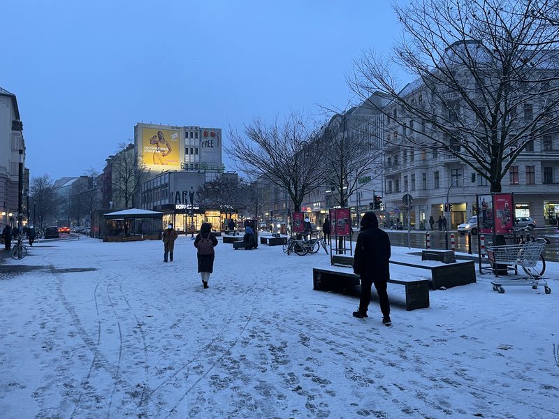 Alfred-Scholz-Platz Neukölln. Eine leichte Schneedecke, Einige Menschen sind zu Fuß unterwegs. Abendlicht eines trüben Tages. Im HIntergrund ein beleuchtetes Werbeplakat.