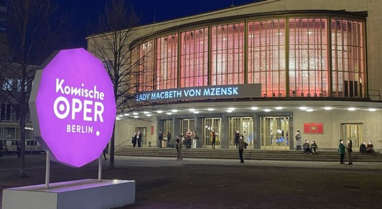 Blick auf das Schillertheater zur Blauen Stunde kurz vor Vorstellungsbeginn. Im Vordergrund das "Komische Oper" Schild, die Ankündigung von Lady Macbeth von Mzensk, das Gebäude von innen beleuchtet, Menschen warten