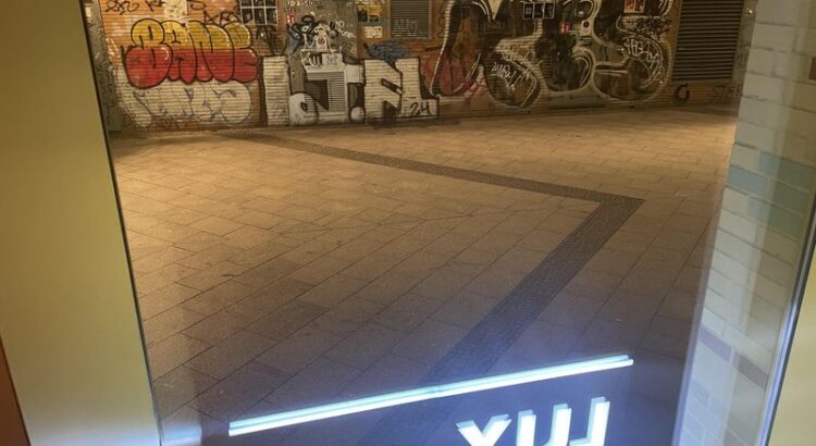 Blick aus dem Delphi Lux Kino. Vor dem Fenster eine abendlich beleuchtete Wand mit Graffitti und ein schmaler Weg. Im Fenster spiegelt sich der Schriftzug "Lux"