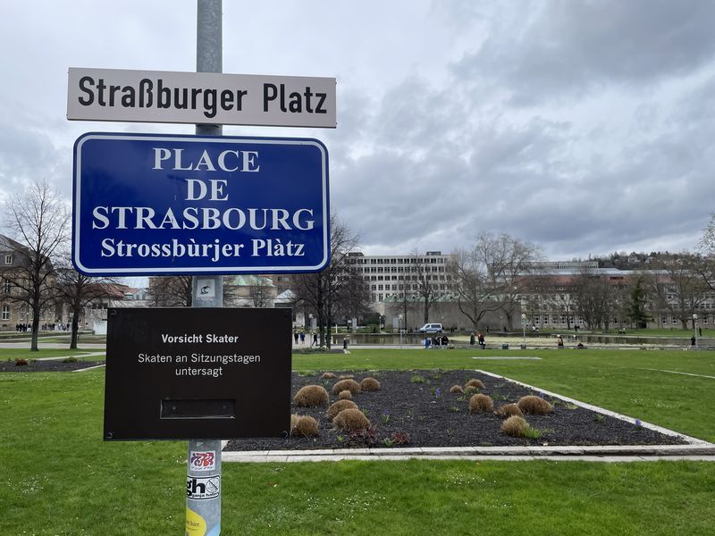 Bild aus dem Stuttharter Schlossgarten. Im Hintegrund Park davor ein halbleeres gepflegtes Bild. Im Vordergrund Bilder "Straßburger Platz / Place des Strasbourg/ Strossburjer Platz" und "Vorsicht Skater. Skaten an Sitzungstagen untersagt"