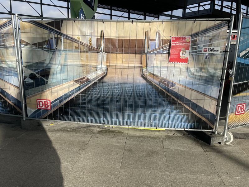 Blick auf einen Bauzaun, hinter dem das brandneue Folienbild einer Rolltreppe zu sehen ist.,