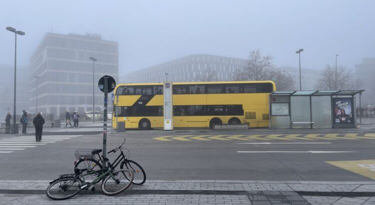 Gelber Doppeldeckerbus in der Nebellandschaft des Bahnhofsvorplatzes. Im Vordergrund zwei Fahrräder, halb umgekippt, halb von ihrem Schloss an einem Schild stabilisiert,