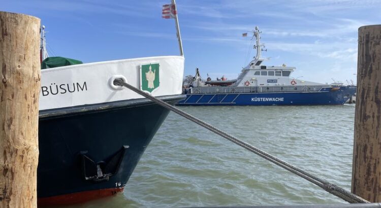Blick ins Hafenbecken Büsum. Links ein Bug auf dem "Büsum" steht zusammen mit einem Neptun.Wappen, weiter hinten ein Schiff der Küstenwache,