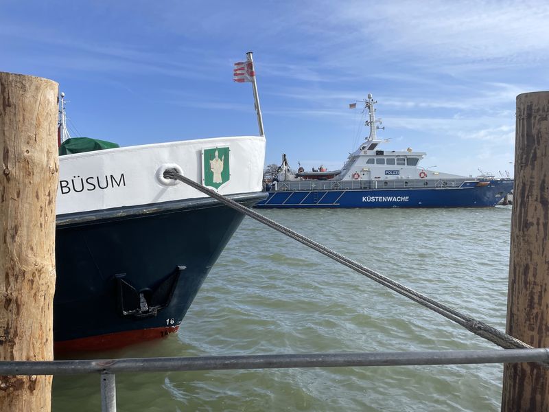 Blick ins Hafenbecken Büsum. Links ein Bug auf dem "Büsum" steht zusammen mit einem Neptun.Wappen, weiter hinten ein Schiff der Küstenwache,