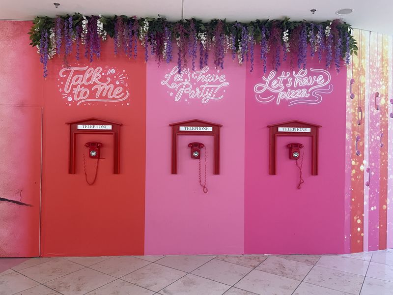 Wand in einem Einkaufszentrum in drei Schattierungen des Rosas. An jeder hängt ein altmodisches Wandtelefon in rot. Darüber die Überschriften, pseudo-neon "Talk to me." "LEts's have party" "Let's have Pizza"